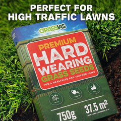 Grassias Premium Hard Wearing Grass Seeds 750g box sitting on green grass and soil. The box features a red label reading 'Premium Hard Wearing Grass Seeds — For a Healthier and Thicker Lawn' with three icons for Spread, Water, and Grow. Coverage listed as 37.5m² for overseeding and 21m² for new lawns. Text overlay reads 'Perfect for High Traffic Lawns.'